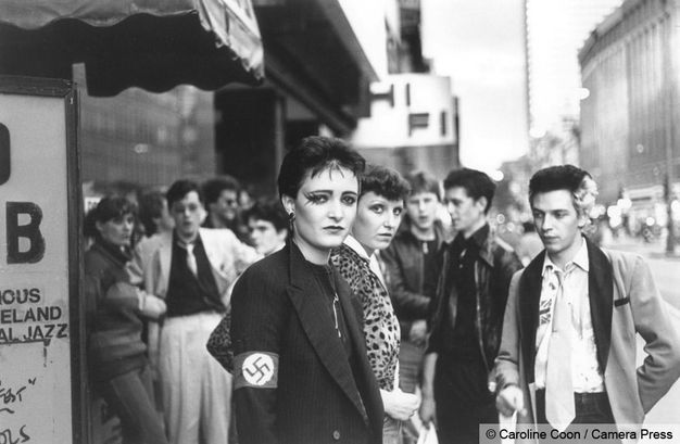 Black and white photograph. A young woman with short hair and elaborate eye liner stands in front of a crowd, many of whom look at us, in front of a club. She wears a black blazer with a swastika band on one arm. 
