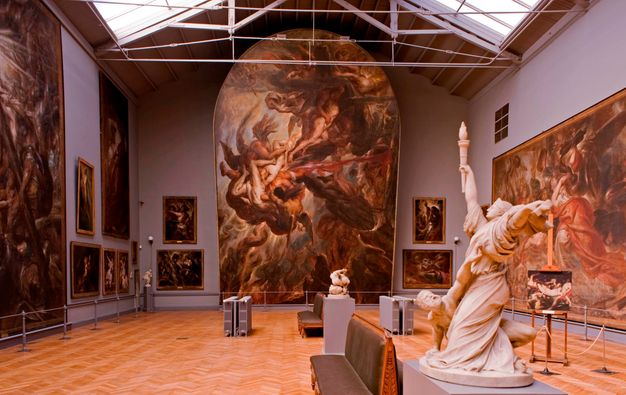 Dynamic paintings of people hang on grey walls under skylights. The arched top of the painting across from us reaches into the peaked ceiling, and a work to our left goes from floor to ceiling. Other works are rather large and some measure a few feet across.